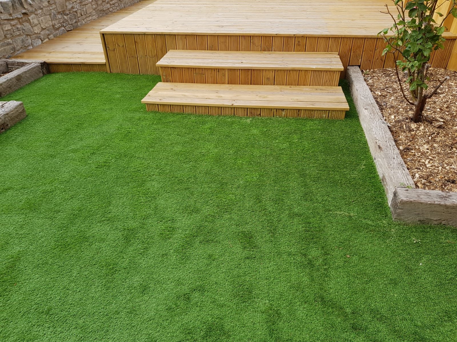Artificial grass fitted beside timber deck steps and stone-edged planting beds
